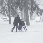 Old man slowly and hard pushing woman in wheelchair in winter day. Heavy snow storm in tree park.