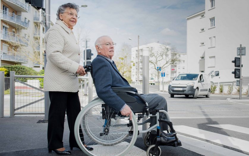 Elderly lady pushing husband in wheelchair across road
