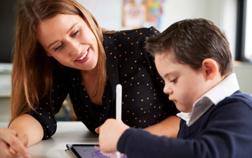 Young female teacher sitting at desk with a Down syndrome schoolboy using a tablet computer