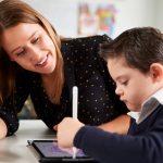 Young female teacher sitting at desk with a Down syndrome schoolboy using a tablet computer