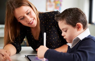 Young female teacher sitting at desk with a Down syndrome schoolboy using a tablet computer