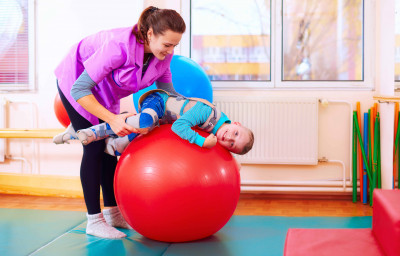 cute kid with disability has musculoskeletal therapy by doing exercises in body fixing belts on fit ball