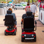 elderly Couple ride along the sidewalk to an electric wheelchair.