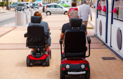elderly Couple ride along the sidewalk to an electric wheelchair.