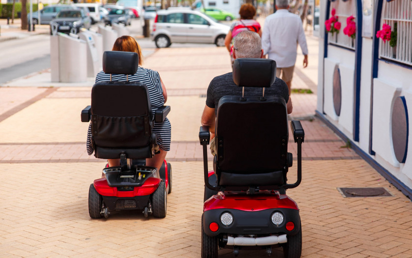 elderly Couple ride along the sidewalk to an electric wheelchair.