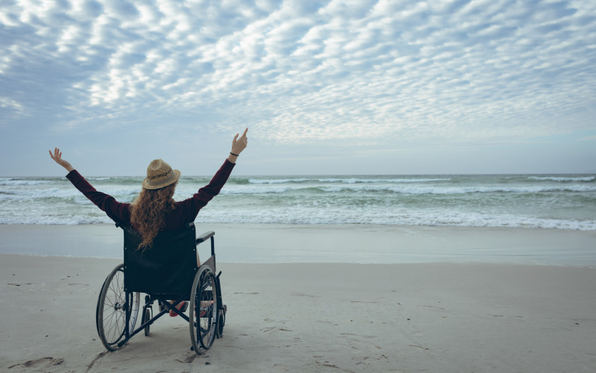 Young disabled woman sitting with arms in the air in wheelchair at beach