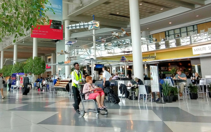 A woman in a wheelchair being pushed through a busy airport main concourse by an attendant to get to her next gate and catch her flight.
