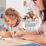 Close-up of a child with autism and teacher by a table drawing with crayons