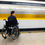 Back view of man in his electric wheelchair at underground platform waiting for train with motion blur of passing train in the background