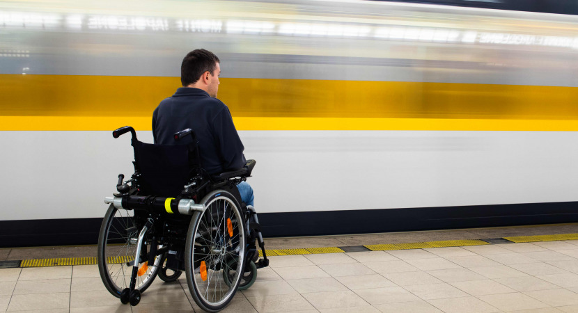 Back view of man in his electric wheelchair at underground platform waiting for train with motion blur of passing train in the background