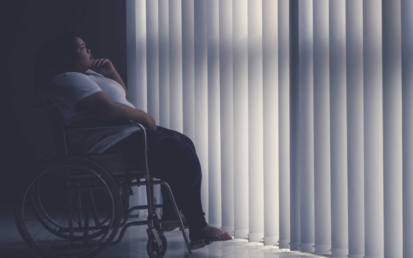 Side view of obese woman looks pensive while sitting in the wheelchair by the window.