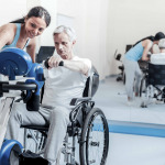 Old man exercising on a training device and sitting in a wheelchair while a smiling woman standing near him