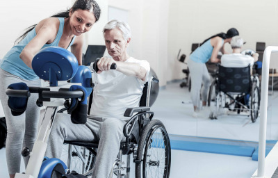 Old man exercising on a training device and sitting in a wheelchair while a smiling woman standing near him