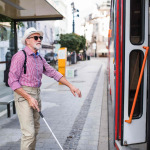 A senior blind man with white cane getting on public transport in city.
