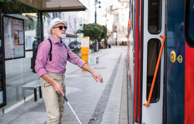 A senior blind man with white cane getting on public transport in city.