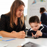 Young female teacher working with a Down syndrome schoolboy sitting at desk using a tablet computer