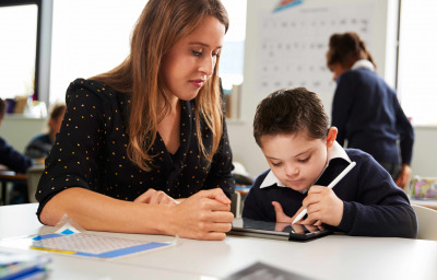 Young female teacher working with a Down syndrome schoolboy sitting at desk using a tablet computer