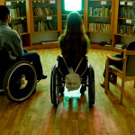 Two people in wheelchairs and one in a chair in front of a TV in the library