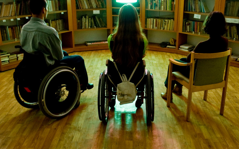 Two people in wheelchairs and one in a chair in front of a TV in the library
