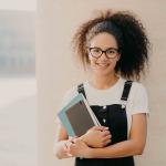 curly haired female student wears white casual t shirt and overalls, holds textbook