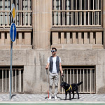 Young blind man with white cane and guide dog standing on pavement in city