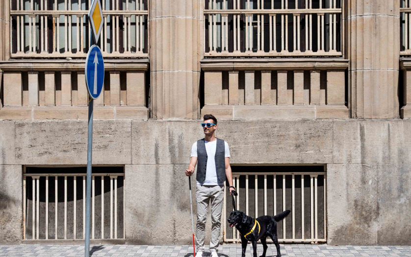 Young blind man with white cane and guide dog standing on pavement in city