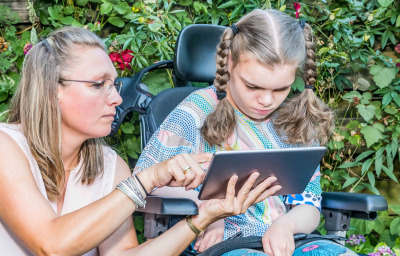 A child in a wheelchair sitting outside looking at a tablet together with a care worker