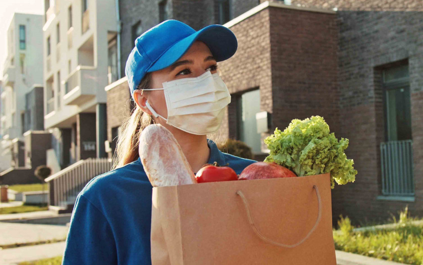 Close up portrait young courier woman holding food craft bag. Modern safety food delivery service. A delivery woman wearing medical mask.