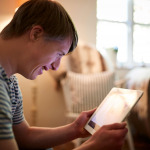 Young Downs Syndrome Man Sitting On Sofa Using Digital Tablet At Home