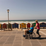 Side view of disabled man being pushed on wheelchair by senior woman at beach on a sunny day