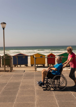 Side view of disabled man being pushed on wheelchair by senior woman at beach on a sunny day