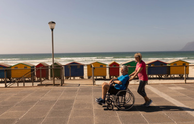 Side view of disabled man being pushed on wheelchair by senior woman at beach on a sunny day