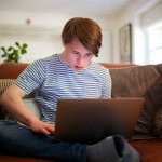 Young Downs Syndrome Man Sitting On Sofa Using Laptop At Home