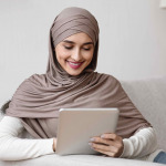 Smiling arabic girl in hijab using digital tablet while relaxing on couch at home