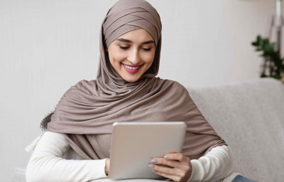 Smiling arabic girl in hijab using digital tablet while relaxing on couch at home
