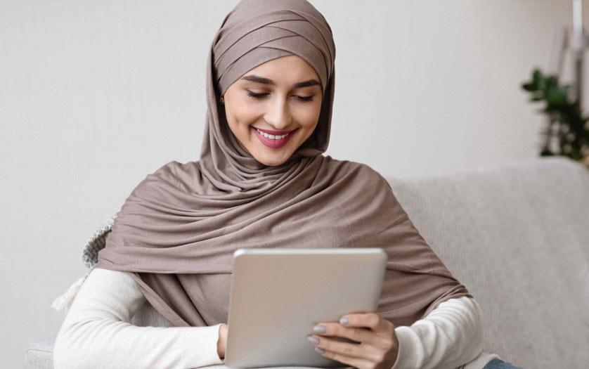 Smiling arabic girl in hijab using digital tablet while relaxing on couch at home