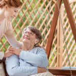An elderly female sitting on a patio swing, Professional caretaker standing next to the woman.