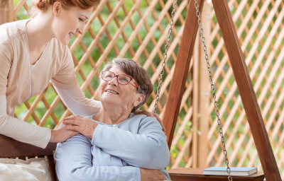 An elderly female sitting on a patio swing, Professional caretaker standing next to the woman.