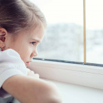 one sad little girl sitting near the window at the day time.