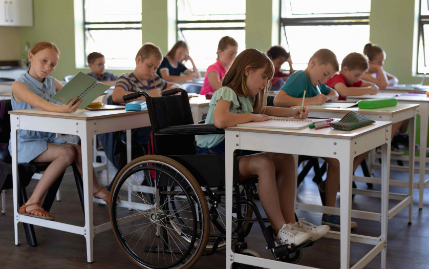 Side view of a diverse group of schoolchildren sitting at desks in classroom, with one schoolgirl sitting in a wheelchair