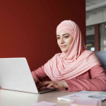 Portrait of young businesswoman wearing headscarf using laptop at desk while working in office against red wall