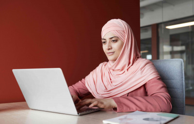 Portrait of young businesswoman wearing headscarf using laptop at desk while working in office against red wall