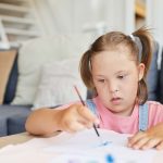 Little girl with down syndrome learning to paint with paints at the table at home