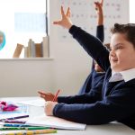 Schoolboy with Down syndrome sitting at a desk raising his hand in a primary school class, close up, side view