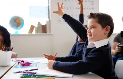Schoolboy with Down syndrome sitting at a desk raising his hand in a primary school class, close up, side view