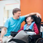 Father talking with son sitting in wheelchair while waiting in doctor s office, laughing together.