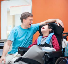 Father talking with son sitting in wheelchair while waiting in doctor s office, laughing together.