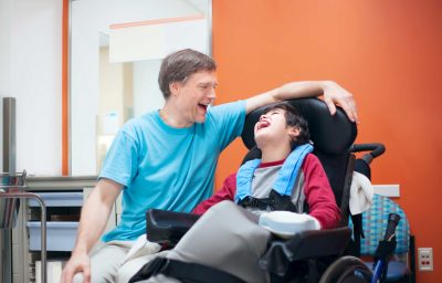 Father talking with son sitting in wheelchair while waiting in doctor s office, laughing together.