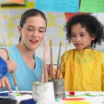 Young teacher assisting cute girl in painting during art class