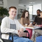 Portrait of smiling red haired boy using wheelchair studying with group of students in college library and looking at camera